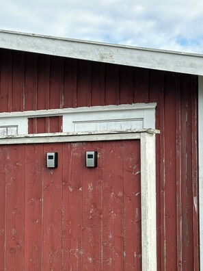 A red barn with a white trim and two small, square, metallic objects mounted on the wall. - Nybovallens Camping (Grönhögen)