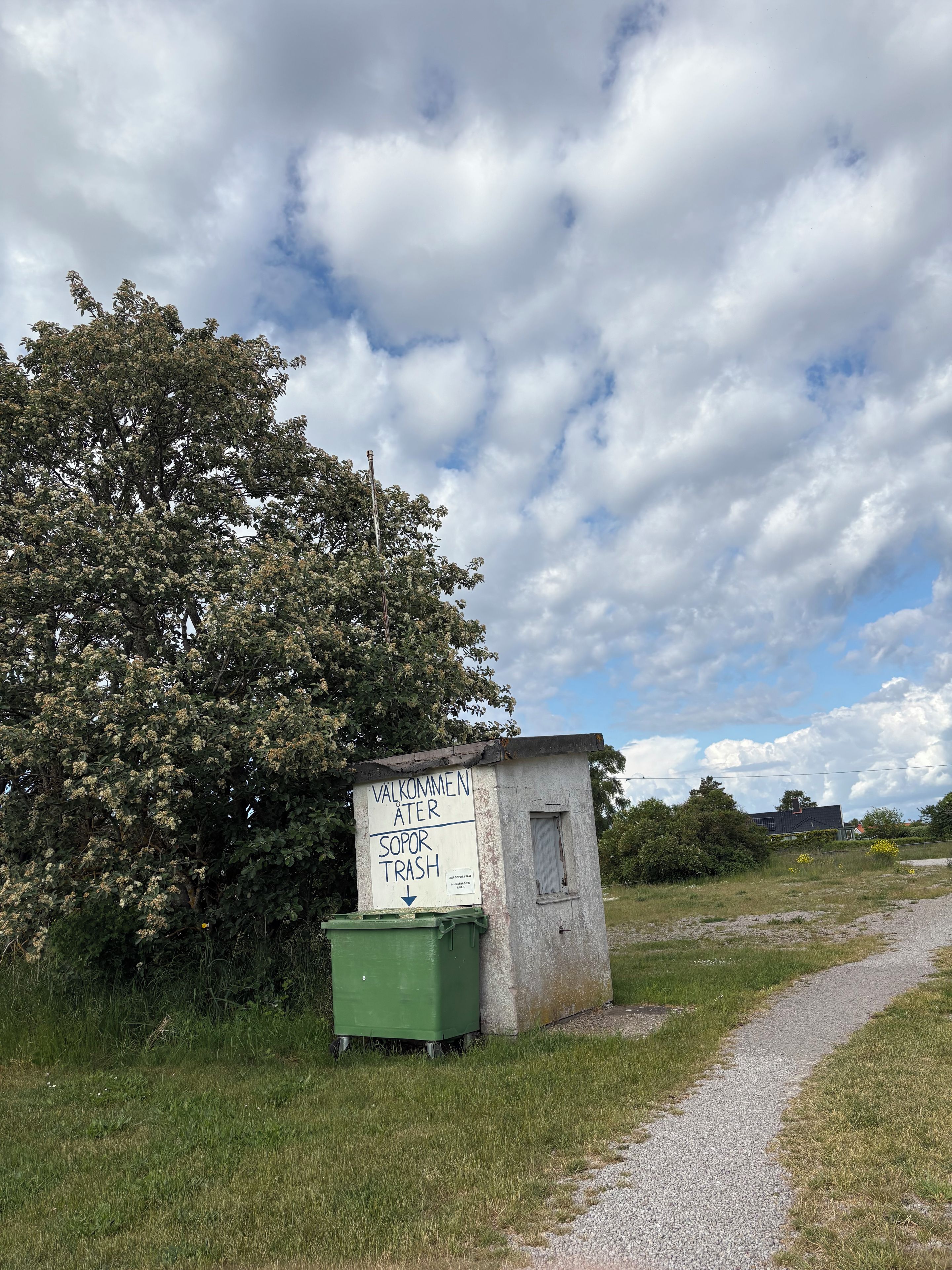 A small, weathered structure with a sign indicating a trash bin, surrounded by grass and a tree.
