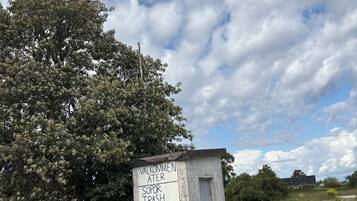 A small, weathered structure with a sign indicating a trash bin, surrounded by grass and a tree.