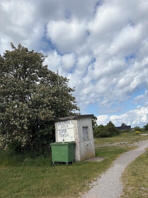 A small, weathered structure with a sign indicating a trash bin, surrounded by grass and a tree. - Nybovallens Camping (Grönhögen)
