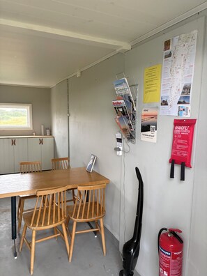 A room with a wooden table and chairs, a window with a view, a wall-mounted fire extinguisher, and various informational posters. - Nybovallens Camping (Grönhögen)