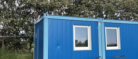 A blue container-style building with two windows and a door, situated in a grassy area with trees in the background.