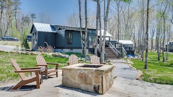 A cabin with a stone fire pit, wooden Adirondack chairs, and a metal staircase leading to a deck.