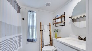 Un baño con inodoro, lavamanos, espejo, ventana con cortinas traslúcidas y toallero.