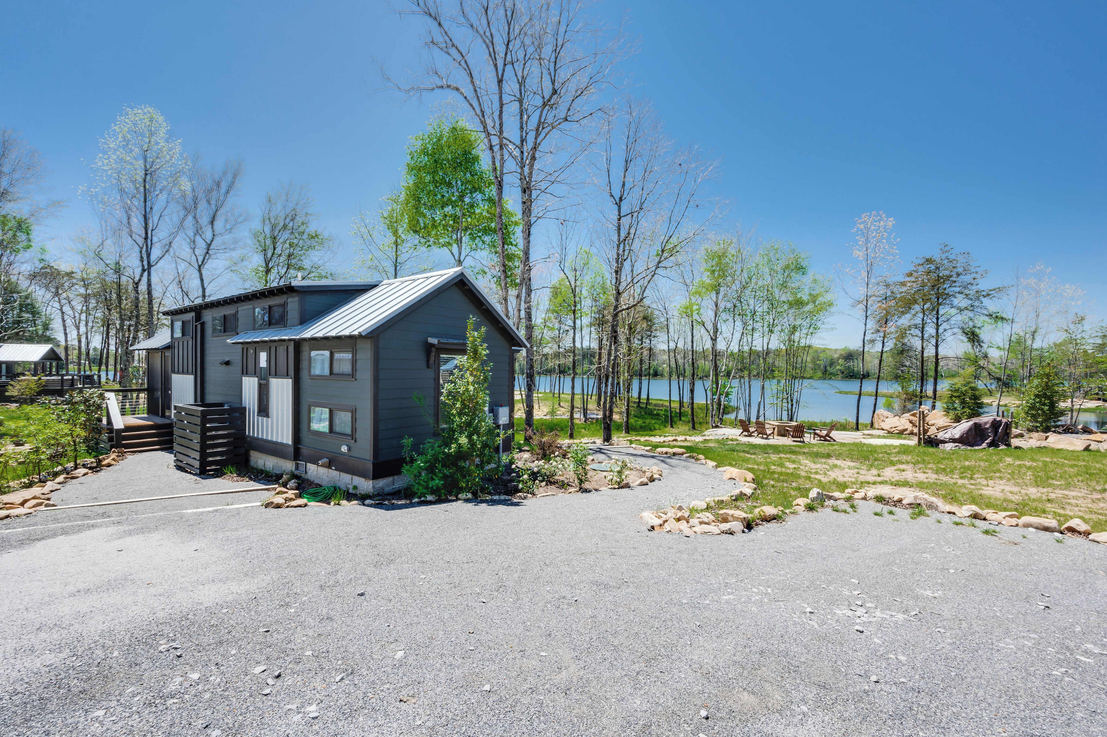 A modern cabin with a gravel driveway, surrounded by trees and a lake.