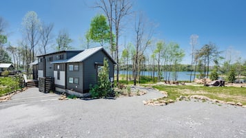 A modern cabin with a gravel driveway, surrounded by trees and a lake.