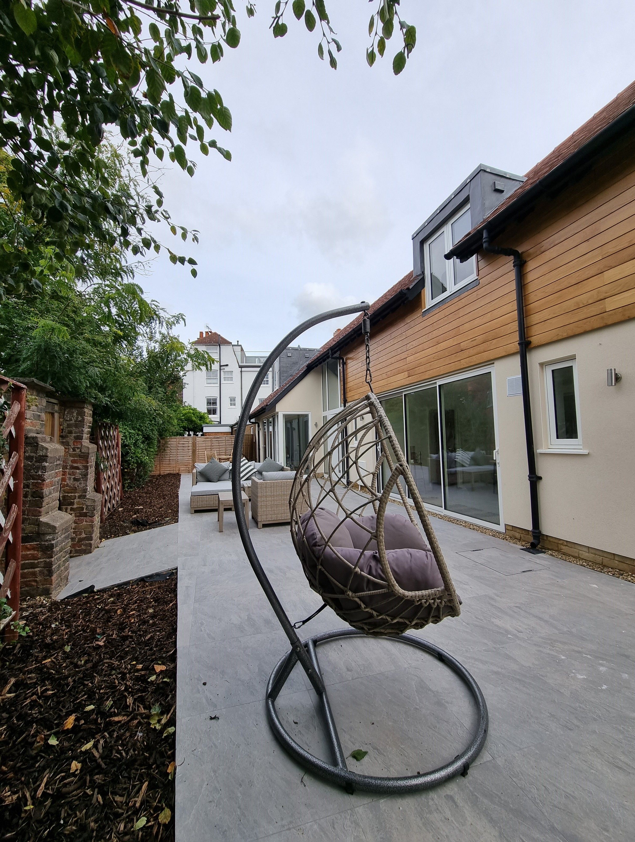 A hanging chair with a cushion, a brick wall, and a building with large windows.