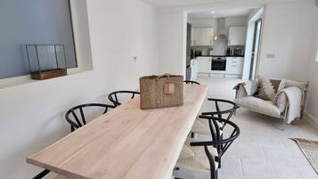 A modern dining area with a wooden table and black chairs, a basket on the table, and a view into a kitchen and living area.