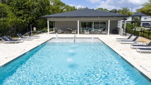 A rectangular swimming pool with clear blue water, surrounded by lounge chairs and a covered seating area with a roof.