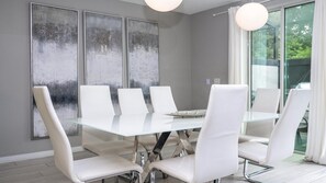 Un comedor moderno con una mesa de vidrio y sillas blancas, grandes cuadros abstractos en la pared, y vistas a través de un amplio ventanal.