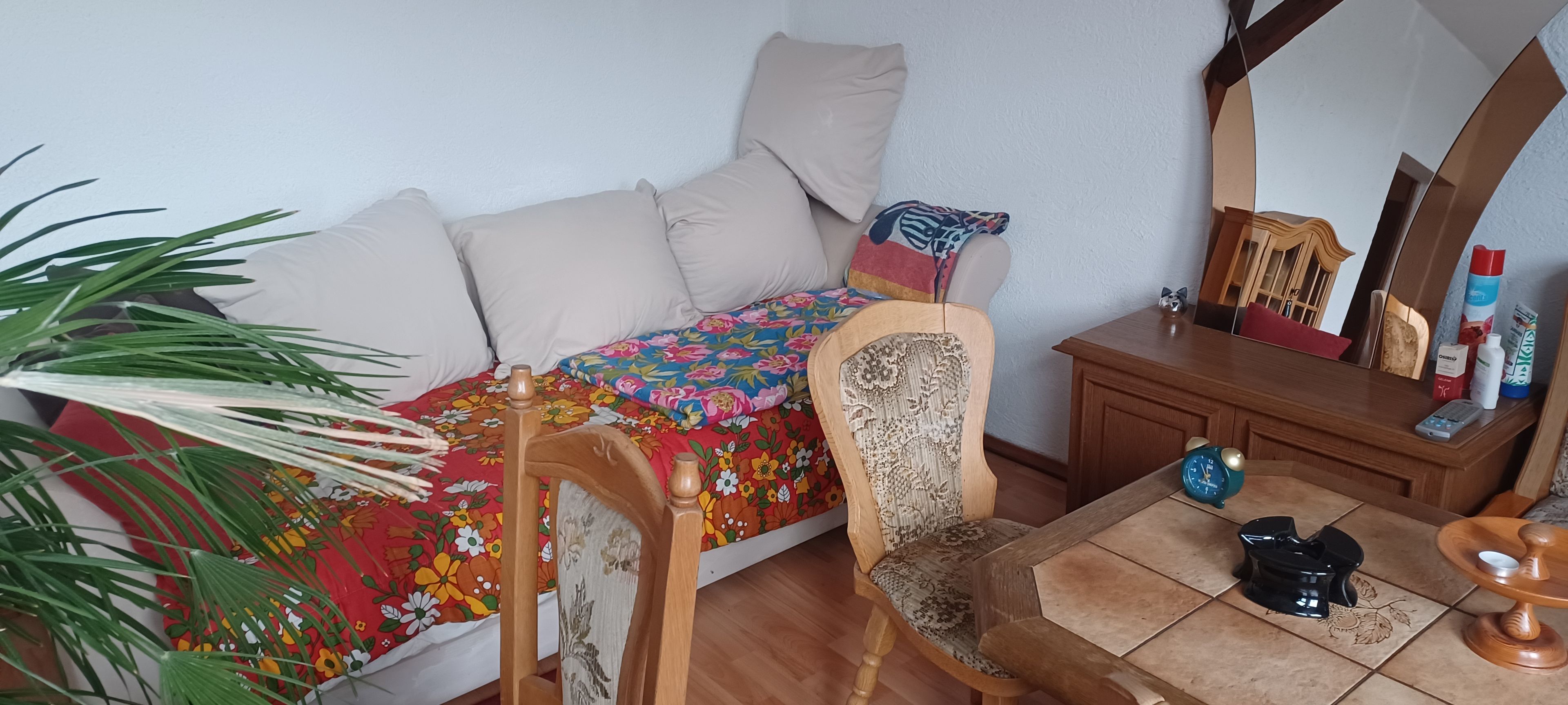 A room with a bed, a chair, a small table, a mirror, and a potted plant.
