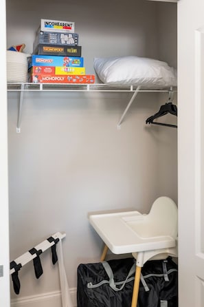 A closet with shelves holding board games, a white bed, a small white table, and a black bag. - Waterfront Retreat + Kayaks + Game Room - Sleeps 6 (Hernando Beach)