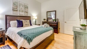Una habitación de hotel con cama, mesita de noche, lámpara, cómoda, espejo y obra de arte en la pared.