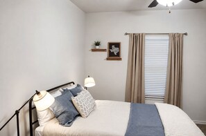 Una cama bien hecha con cabecera, dos lámparas de noche, un pequeño estante con una planta y un cuadro enmarcado, y una ventana con cortinas.