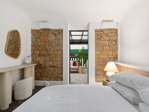 A bedroom with a stone wall, a bed with white bedding, a bedside lamp, and a view through an open door to a balcony with a chair and plant. - Stylish 2-bed House Near Sydney CBD (Sydney)