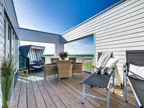 Eine überdachte Terrasse im Freien mit Korbmöbeln, einem Tisch mit Blumenvase und Blick auf das Wasser.