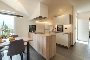 A modern kitchen with a central island, white cabinetry, and a view of a church through a large window. - Holiday Home Lukc (Kobarid)