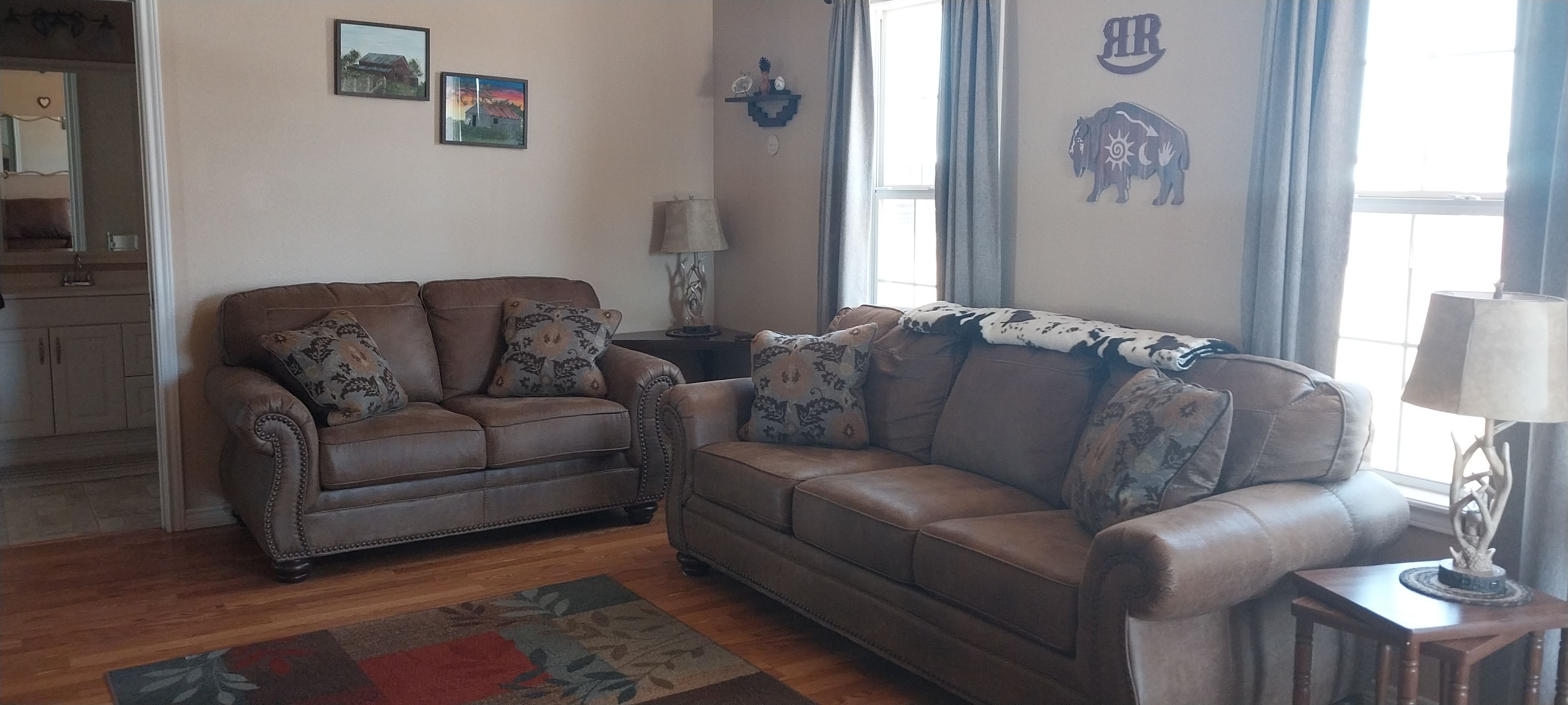 A living room with a brown leather sofa, a wooden coffee table, and a patterned rug. There are two wall-mounted lamps and a window with curtains.