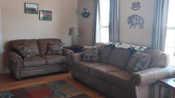 A living room with a brown leather sofa, a wooden coffee table, and a patterned rug. There are two wall-mounted lamps and a window with curtains.