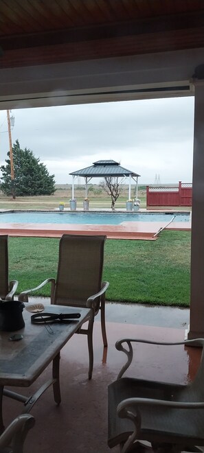 An outdoor patio with a table and chairs, a covered area, and a view of a pool and distant landscape.