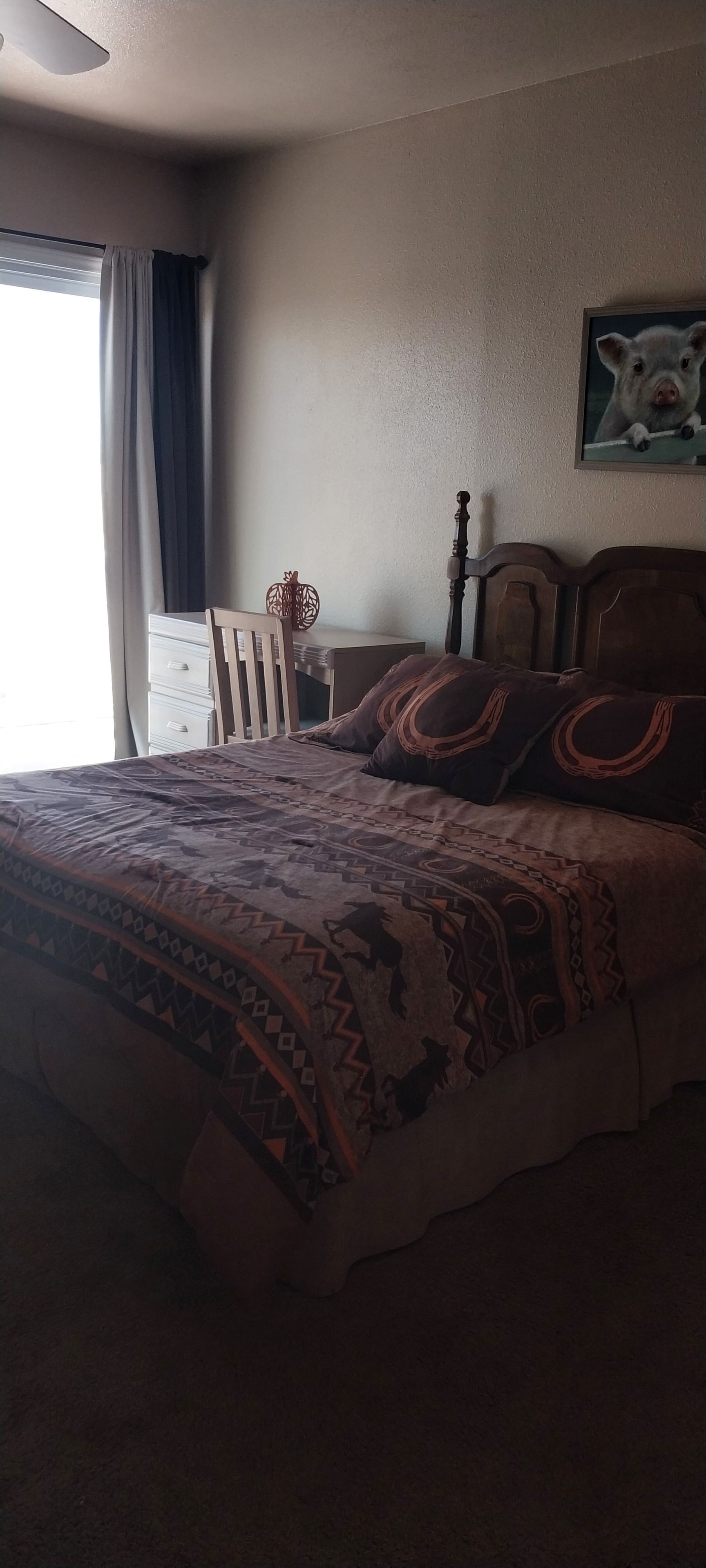 Un dormitorio con cama, mesita de noche, silla, ventana con cortinas e imagen colgada en la pared.