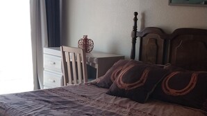 Un dormitorio con cama, mesita de noche, silla, ventana con cortinas e imagen colgada en la pared.