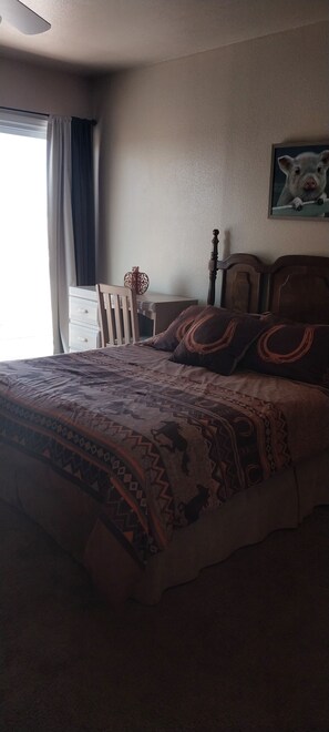 Un dormitorio con cama, mesita de noche, silla, ventana con cortinas e imagen colgada en la pared.