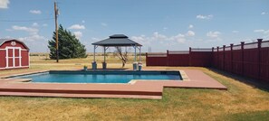 Una piscina rectangular con un pabellón en el centro, rodeada por una cerca roja y un edificio rojo con una puerta blanca.