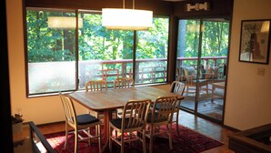 Un coin repas avec une table et des chaises en bois, de grandes fenêtres donnant sur un paysage verdoyant et un tapis rouge à motifs.