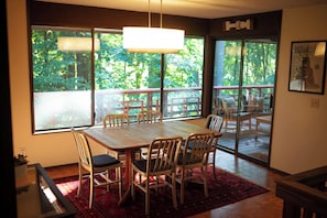 Un comedor con mesa y sillas de madera, ventanas amplias con vista a follaje, y un tapete rojo estampado.