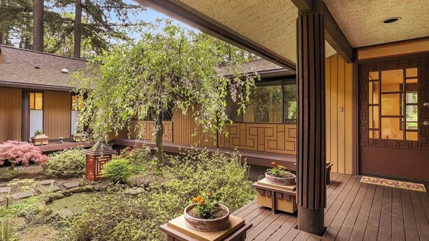 Un espace extérieur abrité, doté d’une terrasse en bois, de plantes en pot et offrant une vue sur une maison avec une porte coulissante de style japonais (shoji).