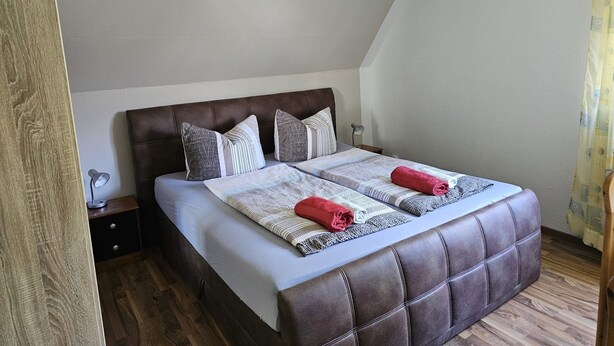 Ein Schlafzimmer mit einem braunen, gepolsterten Kopfteil, einem Bett mit gestreifter Bettwäsche, zwei gerollten Handtüchern, einer Nachttischlampe und einer Hängeleuchte.