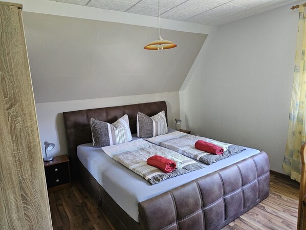Une chambre à coucher avec une tête de lit marron recouverte de tissu, un lit avec une literie à rayures, deux serviettes enroulées, une lampe de chevet et un luminaire suspendu.