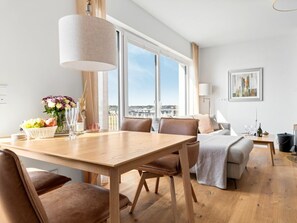 A modern living room with a dining area, large windows, and a view of the city. - Apartment in Olpenitz With Swimming Pool (Kappeln)