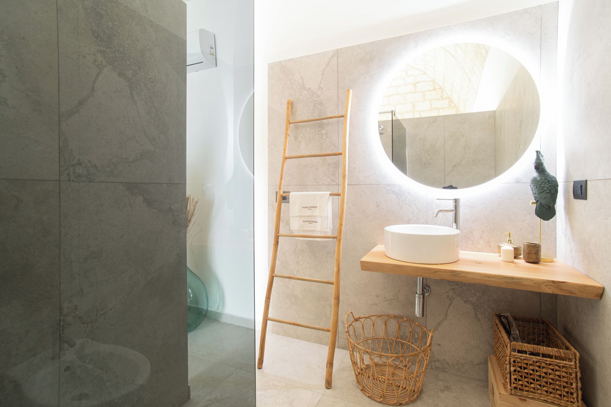 Un bagno con una scala di legno, uno specchio rotondo, un lavandino bianco e un cesto di vimini.