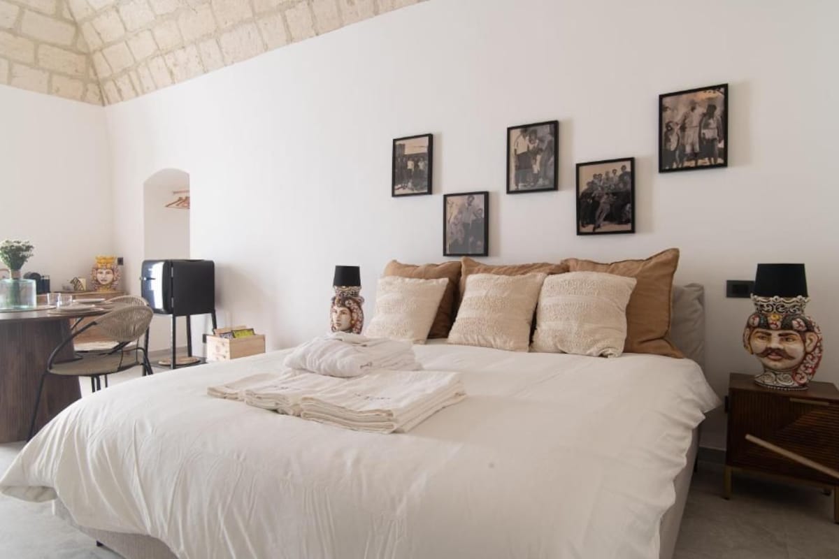 Quarto de hotel com cama, mesas de cabeceira, escrivaninha e uma cadeira. Quadros emoldurados decoram a parede.
