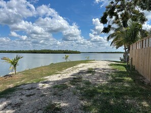 A view of a lake with a sandy beach, a wooden fence, and palm trees. - Waterfront RV Site • Bring Your Own RV (Goodland)