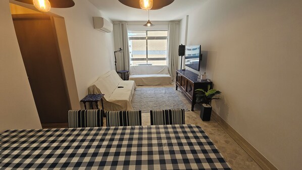 A hotel room with a checkered bedspread, a small table, a sofa, a TV, and a potted plant. - Good Location | Pitangueiras Beach | Wi-fi (Guarujá)