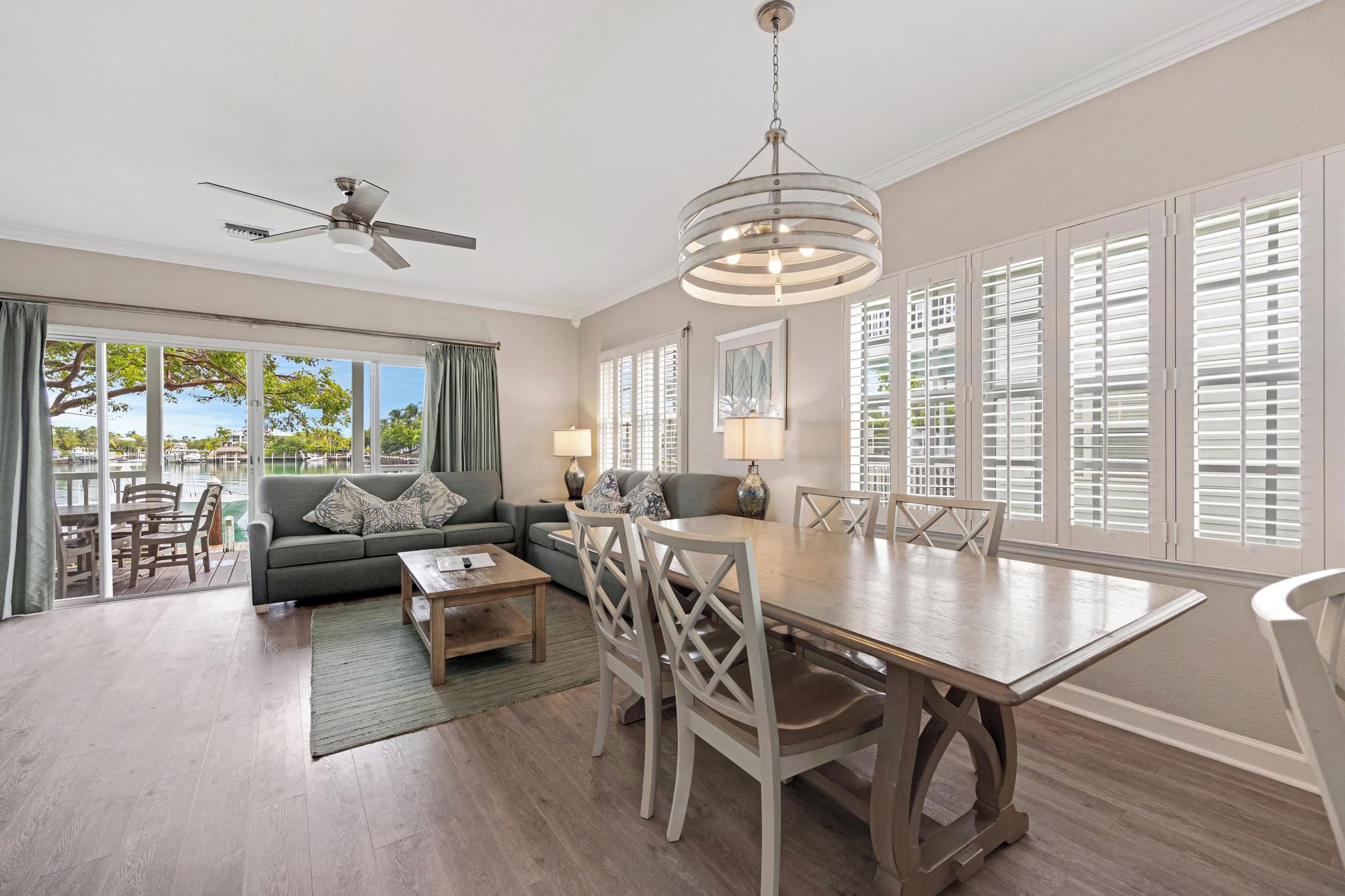 A spacious living room with a dining area, large windows with shutters, a ceiling fan, and a view of the outdoors.