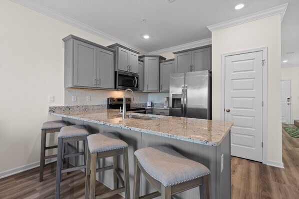 A kitchen with grey cabinets, stainless steel appliances, a granite countertop, and wooden stools. - Luxury Lost Bay Retreat in Orange Beach, AL, 3BR Townhome Near Beach (Orange Beach)