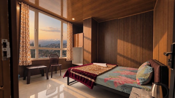 A bedroom with a bed, a desk, and a chair. There is a window with curtains, a view of the outside, and a wooden wall. - DORJE HILL STAY DARJEELING - KANCHENJUNGA VIEW COZY HOMESTAY (Darjeeling)