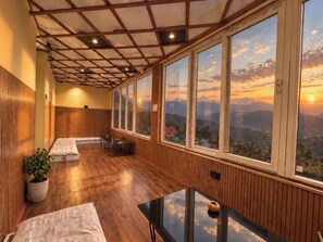 A room with a wooden ceiling, large windows, a bed, a table, and a potted plant.