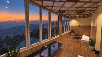 Quarto com uma grande janela com vista para uma paisagem montanhosa, piso de madeira, uma pequena mesa com um vaso e um banco com uma toalha.