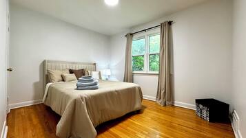 A bedroom with a bed, a window with curtains, a nightstand, and a trash bin.