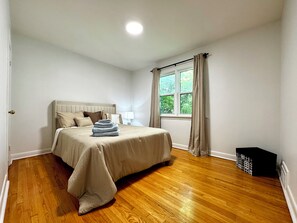 A bedroom with a bed, a window with curtains, a nightstand, and a trash bin. - Newly Furnished Entire House (Toronto)