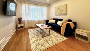 Uma sala de estar com sofá, mesa de centro, TV fixada na parede, janela com cortinas e um quadro.