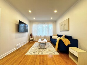 A living room with a sofa, a coffee table, a TV mounted on the wall, and a window with sheer curtains. - Newly Furnished Entire House (Toronto)