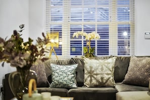 Un salon moderne avec un canapé, des coussins décoratifs, un vase de fleurs et une fenêtre avec des stores.