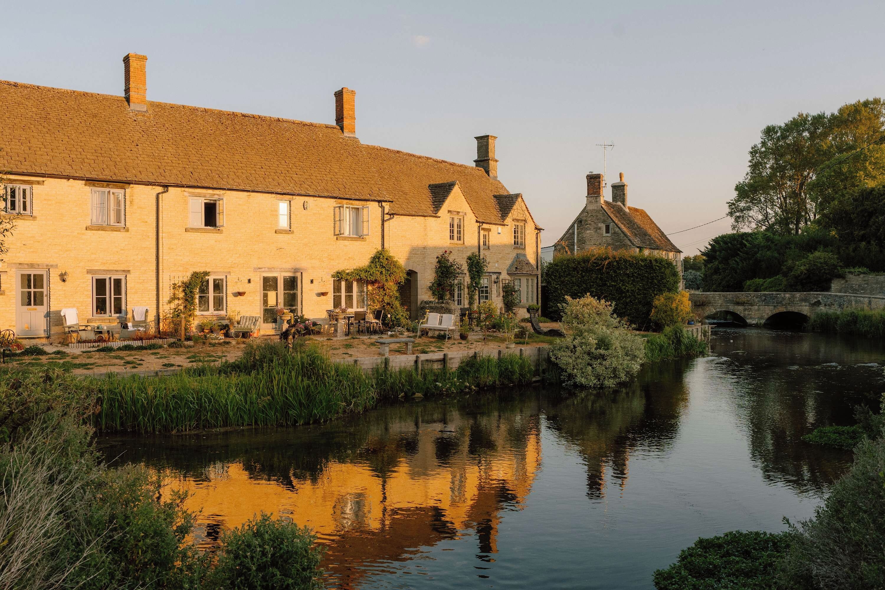Un bâtiment au bord de la rivière, avec une façade en briques, un pont de pierre en arrière-plan et une étendue d’eau calme qui reflète la structure.
