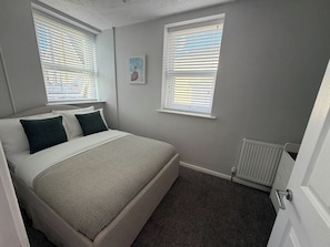 Ein Schlafzimmer mit einem Bett, zwei Fenstern mit Jalousien, einem Heizkörper und einer Tür.
